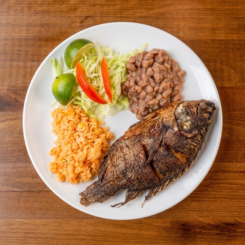 Fried whole fish with rice, beans, shredded lettuce, lime wedges, and red pepper slices.