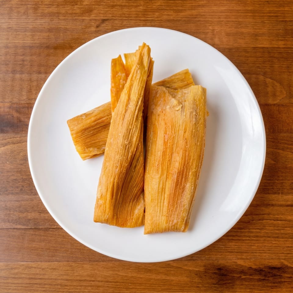 Three golden tamales, still in their corn husk wrappers, arranged on a white plate.