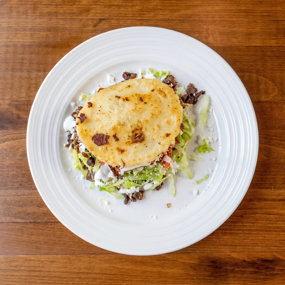Top-down view of a golden-brown arepa filled with meat, lettuce, and white sauce on a white plate.