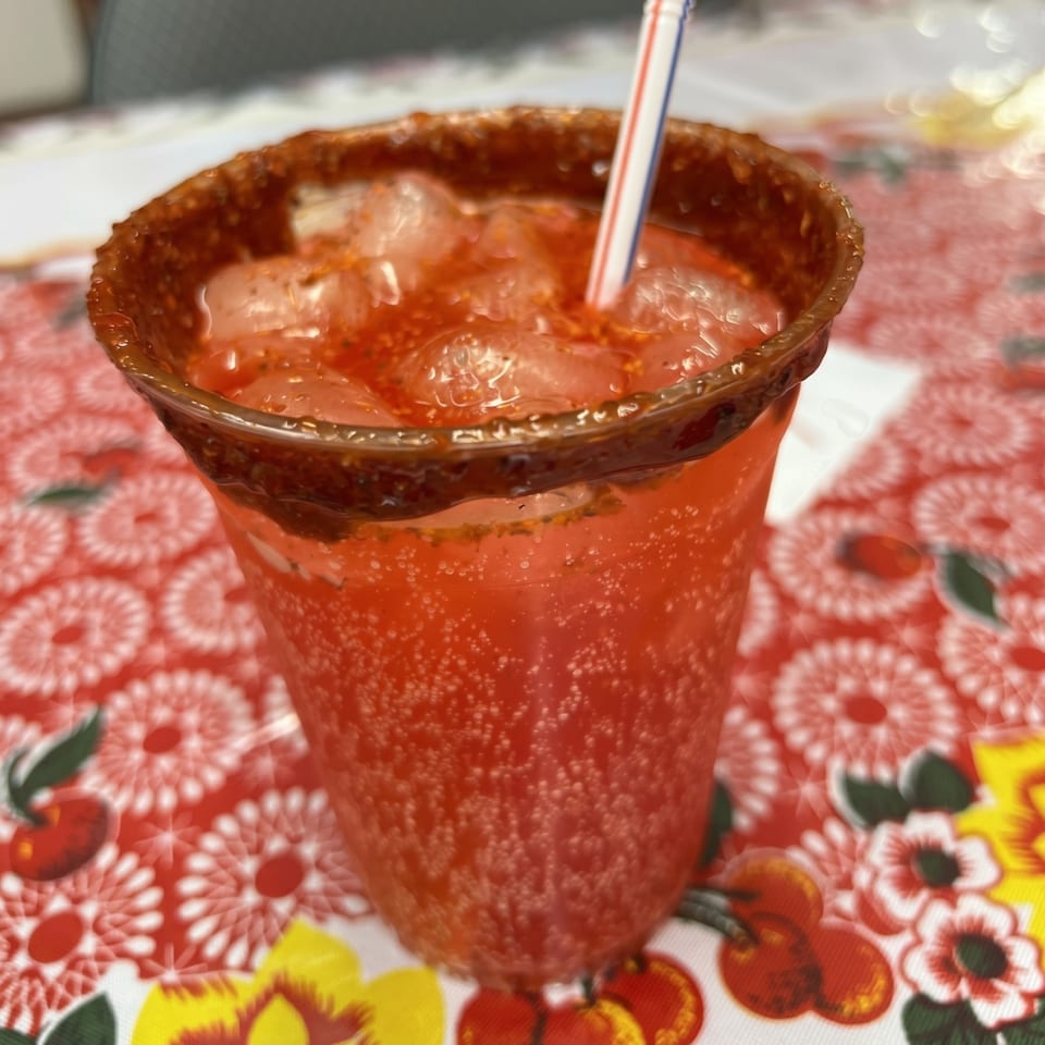 A red, bubbly drink with ice and a straw, rimmed with chili, on a floral tablecloth.