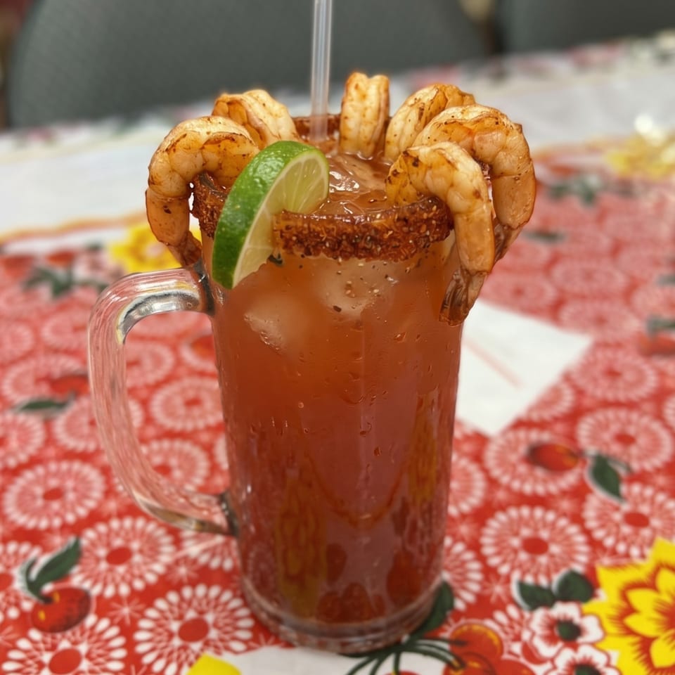 A michelada cocktail with shrimp and a lime wedge on a red floral tablecloth.